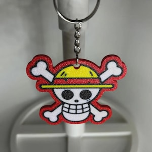 May include: A red, yellow, and white keychain featuring a cartoon skull and crossbones design with a straw hat. The keychain is attached to a silver metal keyring and chain. The design has a black outline.
