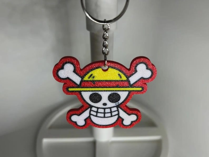 May include: A red, yellow, and white keychain featuring a cartoon skull and crossbones design with a straw hat. The keychain is attached to a silver metal keyring and chain. The design has a black outline.