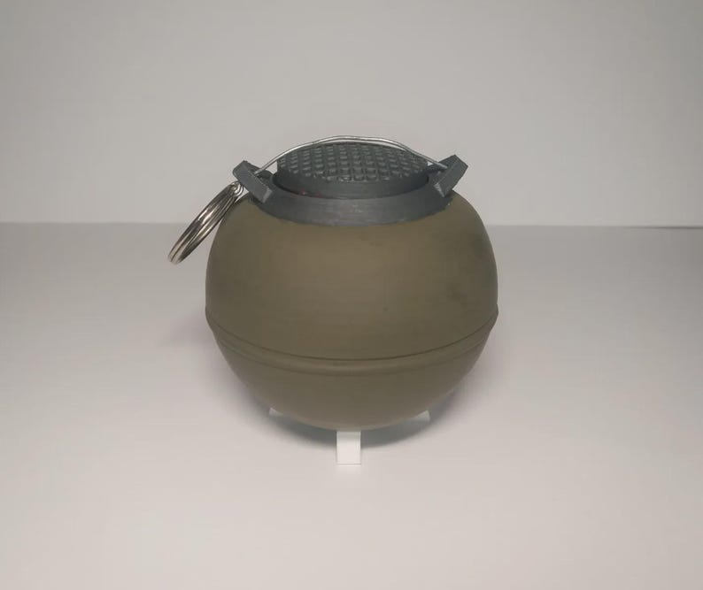 May include: A spherical, olive-drab object with a textured, gray top and a silver ring. The object has a cylindrical shape and is resting on a white stand. The top has a grid-like pattern.