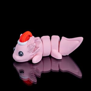 May include: A light pink articulated axolotl figurine featuring a small red and white Santa hat on its head. The creature has large black eyes, external gills, and a segmented body that allows for movement, set against a solid black background.