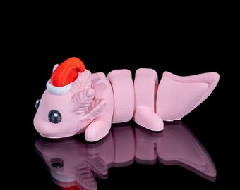 May include: A light pink articulated axolotl figurine featuring a small red and white Santa hat on its head. The creature has large black eyes, external gills, and a segmented body that allows for movement, set against a solid black background.