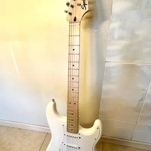 May include: A white Squier Stratocaster electric guitar with a light-coloured maple neck and fretboard. The instrument features a white pickguard, three single-coil pickups, a chrome bridge, and six silver tuning pegs on the headstock.
