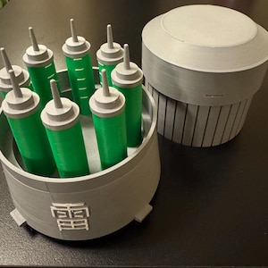 May include: A gray 3D-printed container holding several green cylinders with gray tops and a separate gray cylindrical object with a lid. The container has a Japanese symbol on the side.