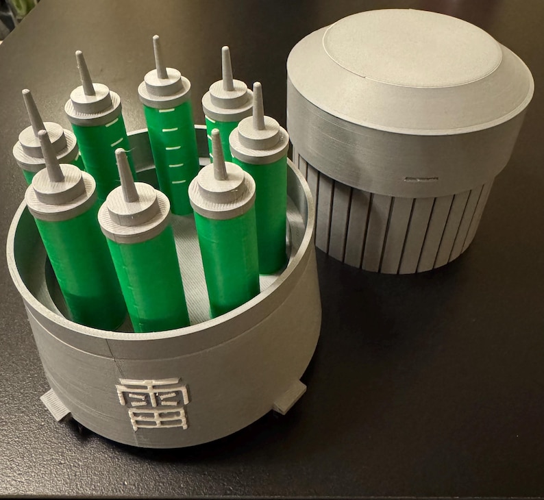 May include: A gray 3D-printed container holding several green cylinders with gray tops and a separate gray cylindrical object with a lid. The container has a Japanese symbol on the side.