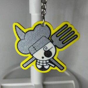 May include: A yellow and gray keychain featuring a cartoon skull with a chef's hat and a fork. The keychain has a yellow outline and a silver chain for attachment. The skull has a black eye and a toothy grin.