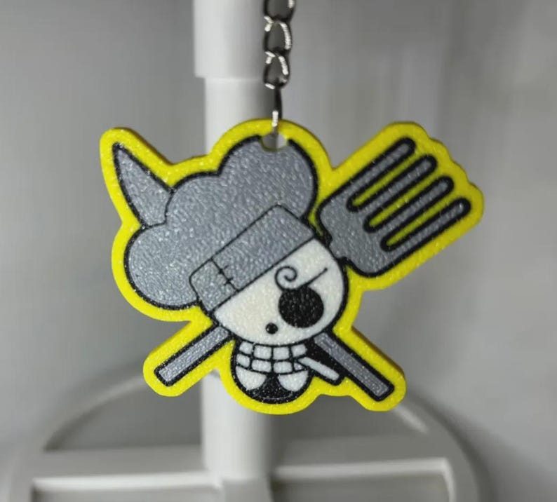 May include: A yellow and gray keychain featuring a cartoon skull with a chef's hat and a fork. The keychain has a yellow outline and a silver chain for attachment. The skull has a black eye and a toothy grin.