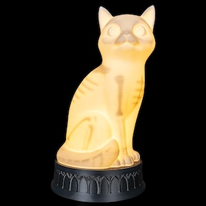 May include: A translucent, off-white cat figurine with an internal skeletal structure visible through the material. The cat is seated in an upright position and is mounted on a circular, black base featuring a repeating arched architectural pattern.