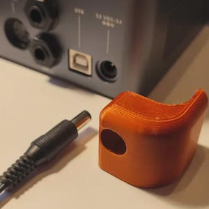 May include: An orange 3D-printed protective cover with a circular opening, next to a black power adapter cable. The cover is designed to fit over a device's power port. The background shows the back of an electronic device.