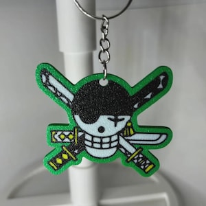 May include: A green and black keychain featuring a cartoon skull with crossed swords. The skull has a black eyepatch and a white face with a grid-like mouth. The swords have yellow and black details. The keychain is attached to a silver metal ring.