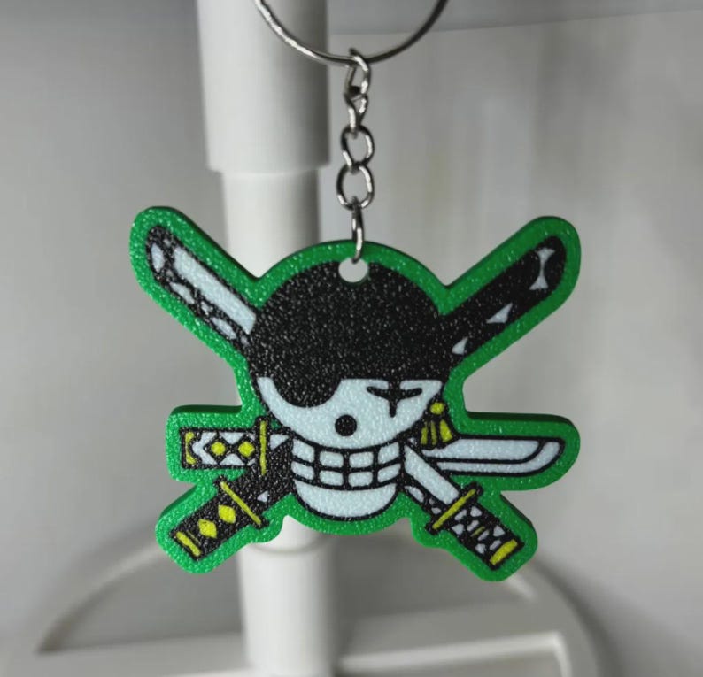 May include: A green and black keychain featuring a cartoon skull with crossed swords. The skull has a black eyepatch and a white face with a grid-like mouth. The swords have yellow and black details. The keychain is attached to a silver metal ring.