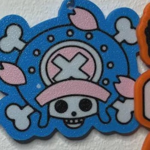 May include: A blue, white, and pink cartoon character pendant. The pendant features a skull with a pink hat and a cross symbol. The design is surrounded by white clouds and pink flower petals. A silver metal loop is at the top.