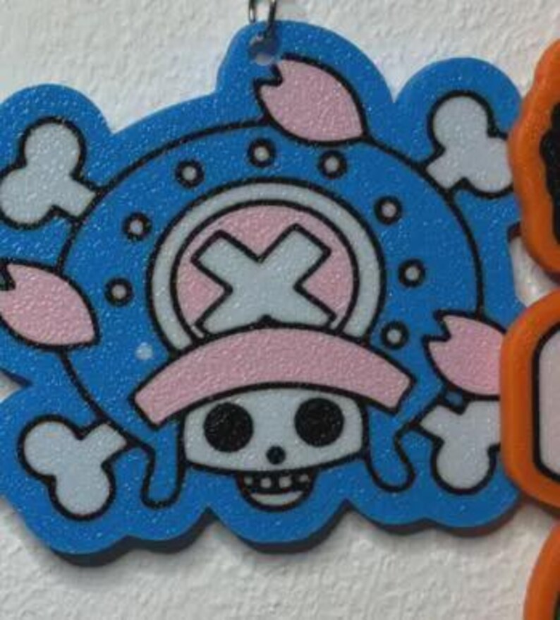 May include: A blue, white, and pink cartoon character pendant. The pendant features a skull with a pink hat and a cross symbol. The design is surrounded by white clouds and pink flower petals. A silver metal loop is at the top.