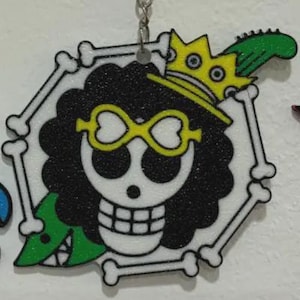 May include: A hexagonal keychain featuring a skull with black curly hair, yellow heart-shaped glasses, and a yellow crown. The design includes a green guitar and is framed by white bones.
