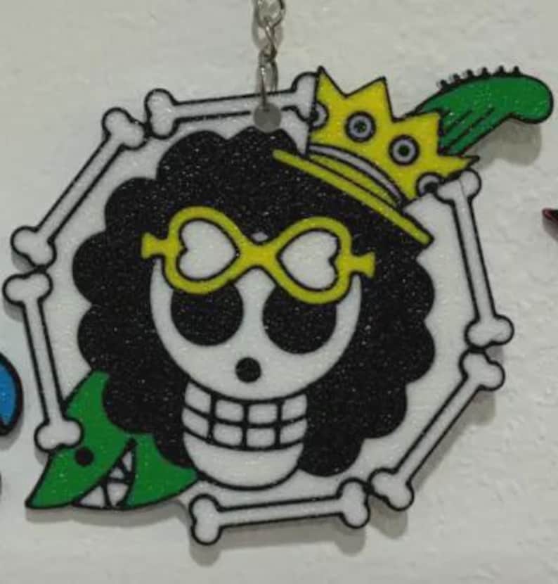 May include: A hexagonal keychain featuring a skull with black curly hair, yellow heart-shaped glasses, and a yellow crown. The design includes a green guitar and is framed by white bones.