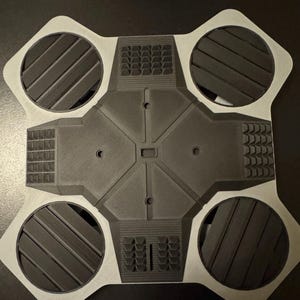 May include: A gray and black geometric object with four circular sections and a central square. The circular sections have angled lines, and the square has a textured surface. The object is likely a component or part of a larger device.