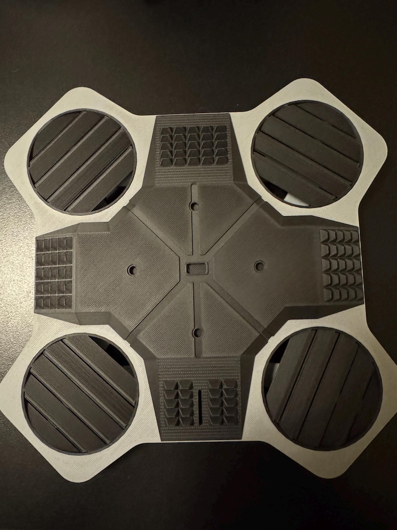 May include: A gray and black geometric object with four circular sections and a central square. The circular sections have angled lines, and the square has a textured surface. The object is likely a component or part of a larger device.