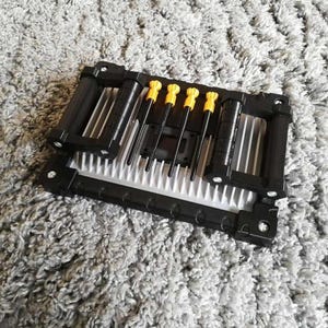 May include: A black and silver computer component with four yellow-handled screwdrivers. The component has a rectangular shape with a series of parallel fins. The screwdrivers are arranged in a row, with their tips pointing towards the center of the component.