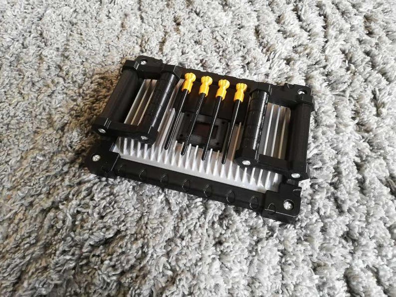 May include: A black and silver computer component with four yellow-handled screwdrivers. The component has a rectangular shape with a series of parallel fins. The screwdrivers are arranged in a row, with their tips pointing towards the center of the component.
