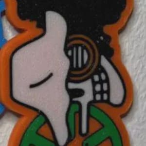 May include: An orange and black graphic of a person with an afro, a white face, and green shapes at the bottom. The design has a small metal loop at the top for hanging. The overall design is a cartoon style.