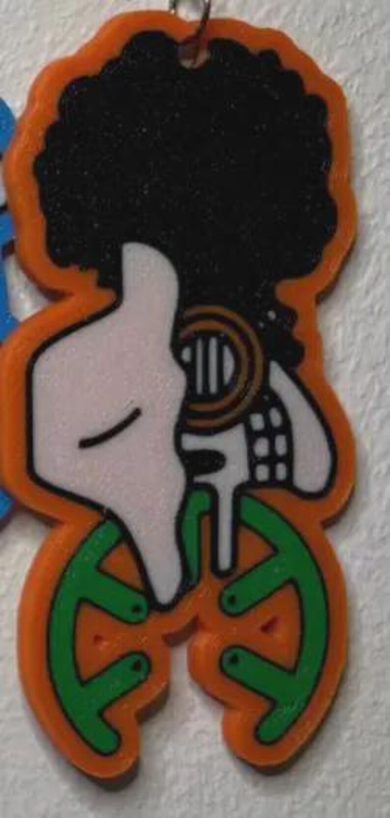 May include: An orange and black graphic of a person with an afro, a white face, and green shapes at the bottom. The design has a small metal loop at the top for hanging. The overall design is a cartoon style.