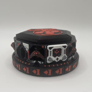 Könnte beinhalten: Eine schwarz-rote dekorative Box mit geometrischem Design. Die Box hat eine kreisförmige Basis mit pfeilartigen Formen und eine Oberseite mit floralem Design. Die Box hat weiße und rote Akzente.