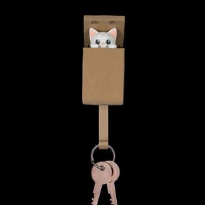 May include: A brown rectangular wall-mounted key holder featuring a small white cat figurine peeking out from the top compartment. A metal ring hangs from the bottom of the holder, securing two silver-colored metal keys in place.