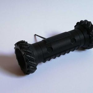 May include: A black 3D-printed object with a cylindrical body and textured ends resembling tires. The object has a series of circular indentations along its length and a thin, black rod extending from the side.