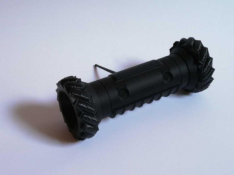May include: A black 3D-printed object with a cylindrical body and textured ends resembling tires. The object has a series of circular indentations along its length and a thin, black rod extending from the side.