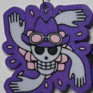 May include: A purple, die-cut charm featuring a skull wearing a hat and goggles, with outstretched arms and a floral design. The charm has a key ring attachment at the top. The design is in white, pink, and black.
