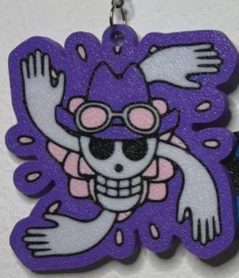May include: A purple, die-cut charm featuring a skull wearing a hat and goggles, with outstretched arms and a floral design. The charm has a key ring attachment at the top. The design is in white, pink, and black.