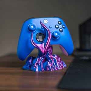 Può includere: Un controller wireless per videogiochi di colore blu brillante appoggiato su un supporto decorativo. Il supporto presenta un design organico e vorticoso con una finitura metallica che alterna tonalità vivaci di viola e blu, sostenendo il controller.