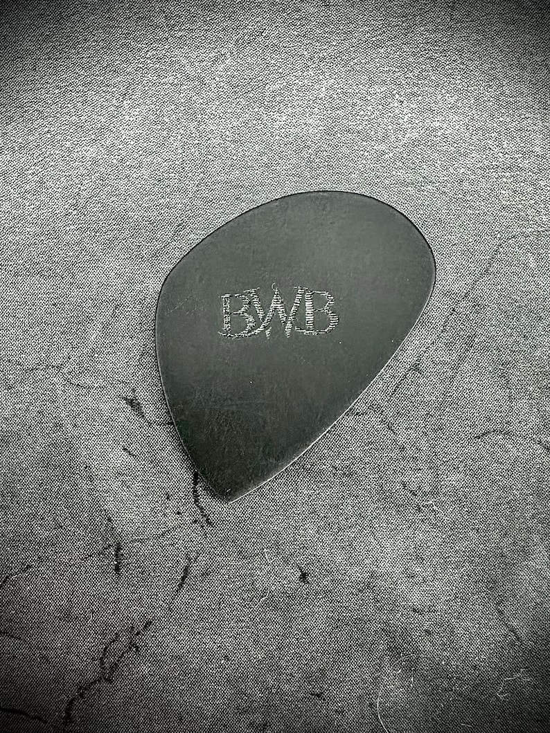 May include: A black guitar pick with the engraved letters "BWB" in the center. The pick has a smooth, rounded shape and is made of a dark material. The background is a textured, dark gray surface.