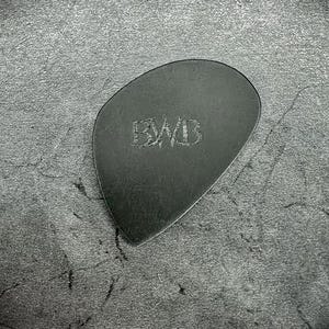 May include: A black guitar plectrum with the engraved letters "BWB" in the centre. The plectrum has a smooth, rounded shape and is made of a dark material. The background is a textured, dark grey surface.