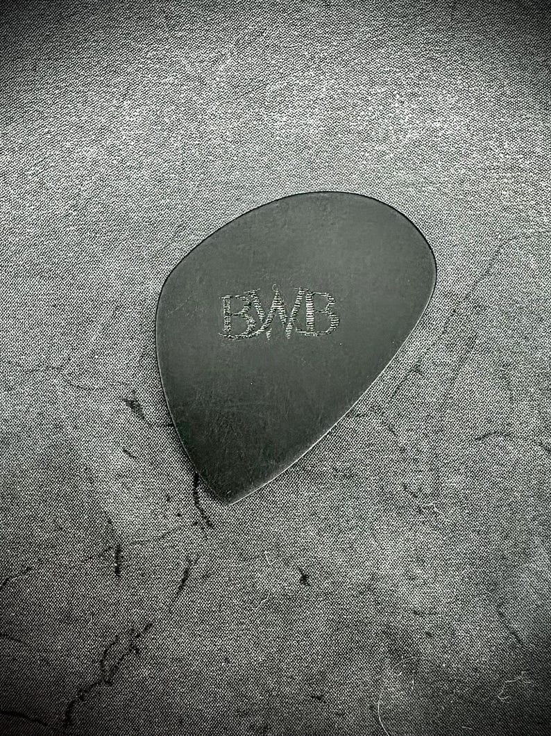 May include: A black guitar plectrum with the engraved letters "BWB" in the centre. The plectrum has a smooth, rounded shape and is made of a dark material. The background is a textured, dark grey surface.