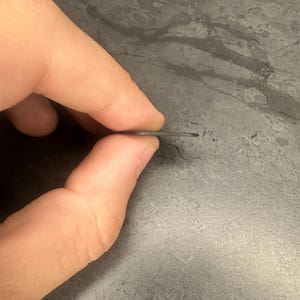 May include: A thin, dark grey metal object is held between fingers. The object appears to be a small, flat piece of metal, possibly a tool or component. The edges are clean and the overall shape is rectangular.