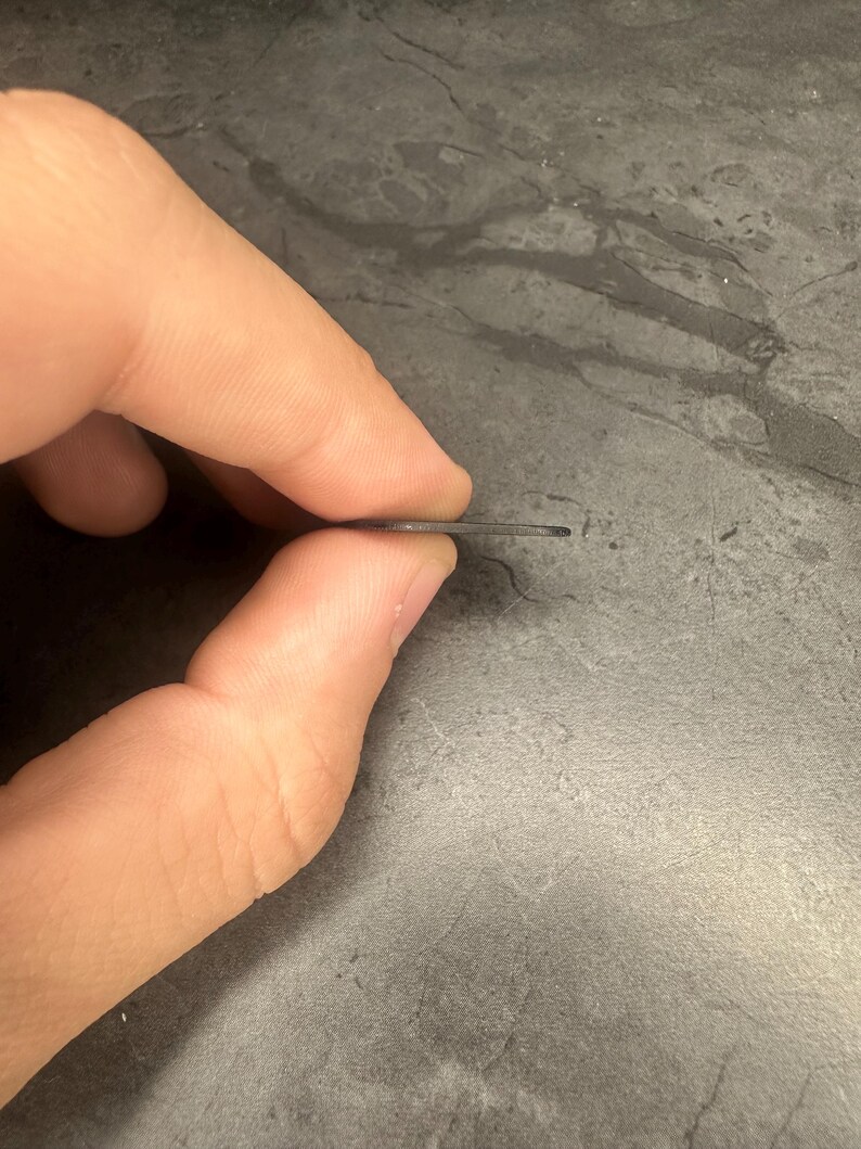 May include: A thin, dark grey metal object is held between fingers. The object appears to be a small, flat piece of metal, possibly a tool or component. The edges are clean and the overall shape is rectangular.