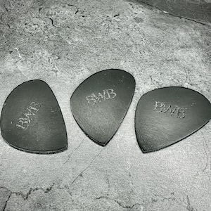 May include: Three black leather guitar picks, each with a distinctive shape and the embossed letters "BWB". They are displayed on a textured grey surface, highlighting their dark colour and design.