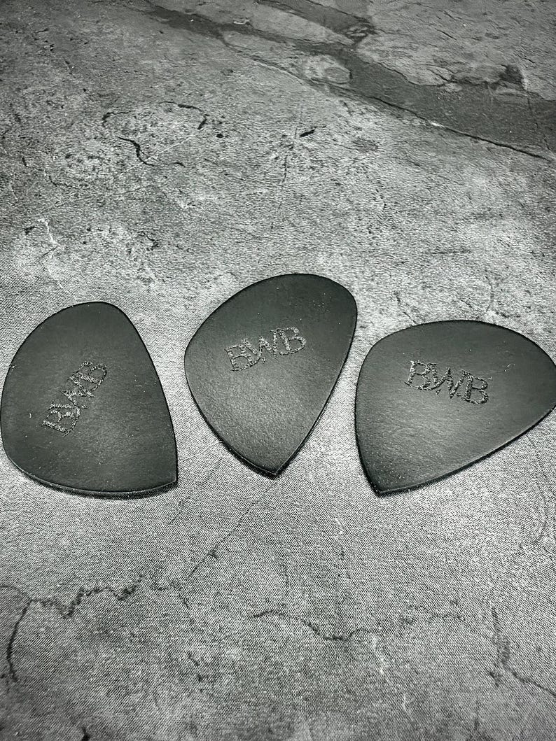 May include: Three black leather guitar picks, each with a distinctive shape and the embossed letters "BWB". They are displayed on a textured grey surface, highlighting their dark colour and design.