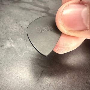 May include: A dark grey guitar plectrum with the embossed letters "PWB". The plectrum is held between fingers, showing its triangular shape and smooth edges. A close-up shot highlights the plectrum's texture and design.