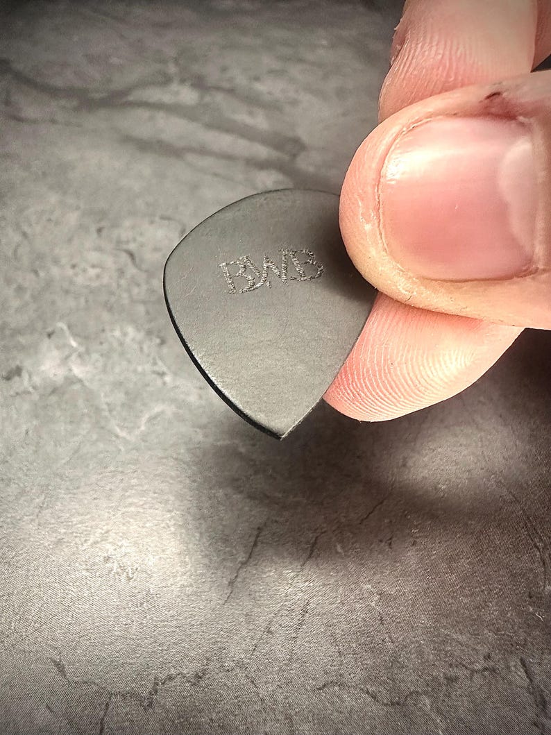 May include: A dark grey guitar plectrum with the embossed letters "PWB". The plectrum is held between fingers, showing its triangular shape and smooth edges. A close-up shot highlights the plectrum's texture and design.
