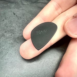 May include: A black guitar plectrum with the embossed letters "BWB". The plectrum is held between fingers, highlighting its shape and texture. The pick has a smooth, matte finish and a rounded triangle shape, commonly used for playing stringed instruments.