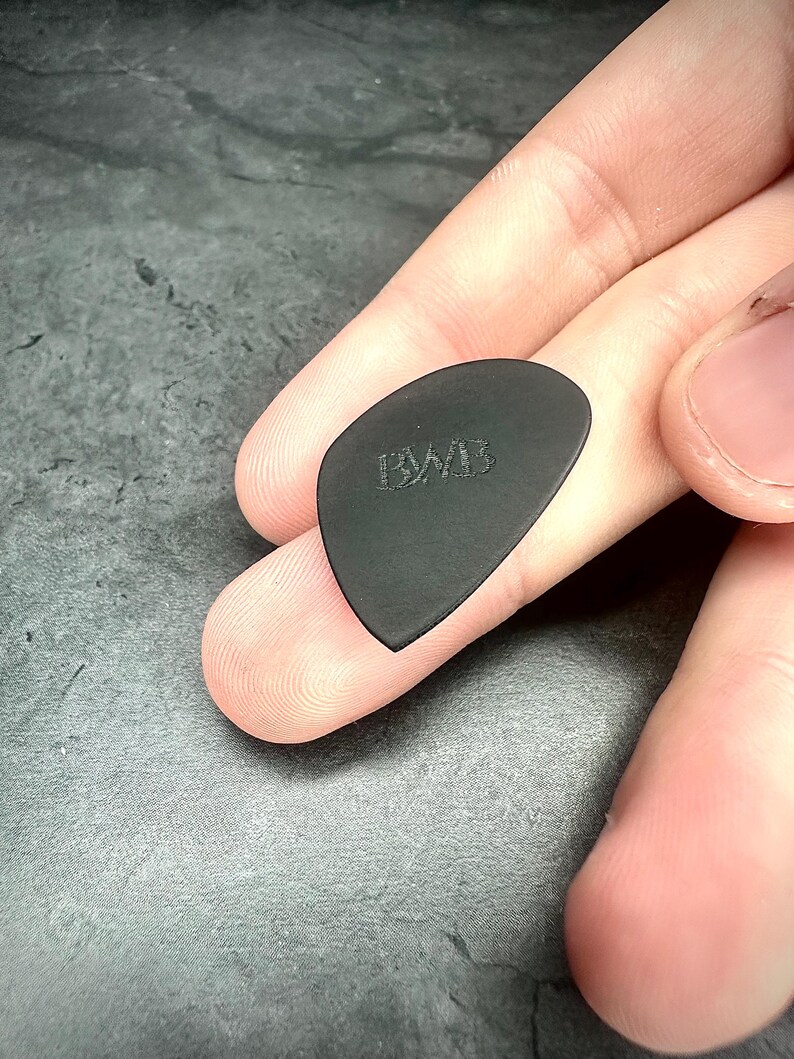 May include: A black guitar plectrum with the embossed letters "BWB". The plectrum is held between fingers, highlighting its shape and texture. The pick has a smooth, matte finish and a rounded triangle shape, commonly used for playing stringed instruments.
