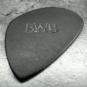 May include: A black guitar plectrum with the embossed letters "BWB". The plectrum has a smooth surface and a rounded triangular shape. The background is a textured, grey surface.