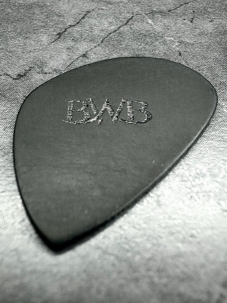 May include: A black guitar plectrum with the embossed letters "BWB". The plectrum has a smooth surface and a rounded triangular shape. The background is a textured, grey surface.