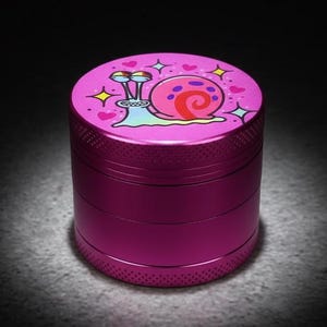 Peut inclure: Un broyeur à herbes cylindrique magenta. Le dessus présente un dessin de l'escargot de dessin animé avec des cœurs et des étoiles. Le broyeur est en métal et a une surface texturée.
