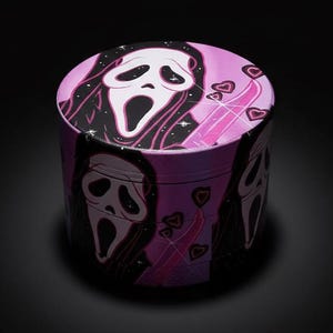 Peut inclure: Un broyeur rond violet avec un motif répété du masque Ghostface de la série de films d'horreur Scream. Le dessus et les côtés présentent le masque, des cœurs roses et une palette de couleurs rose et noire. Le broyeur est posé sur un fond noir.