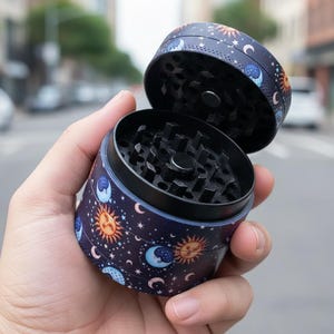 Pode incluir: Um moedor de ervas azul escuro com um design celestial de sóis, luas e estrelas. O moedor está aberto, revelando os dentes de moagem. O design é impresso no exterior.