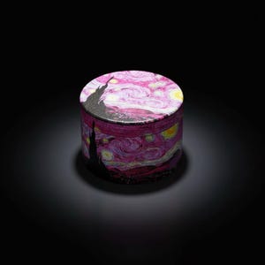 May include: A cylindrical box featuring a print of Van Gogh's "Starry Night" painting. The box has a pink and purple swirling sky design with yellow stars and a black silhouette of a town. The box is set against a black background.