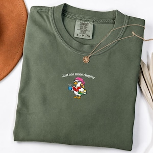 Peut inclure: Un t-shirt à col rond vert olive avec un motif représentant un canard portant un chapeau rose et des lunettes, tenant une tasse bleue et un livre rouge. Le texte "Just one more chapter" est imprimé au-dessus du canard. Étiquette Comfort Colors.
