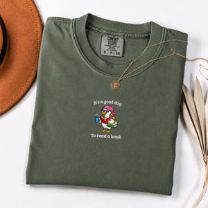 Peut inclure: Un t-shirt à col rond vert olive avec une illustration d'un canard blanc portant un chapeau rose et des lunettes, tenant une tasse bleue et un livre rouge, avec le texte "It's a good day To read a book" imprimé au-dessus et en dessous.